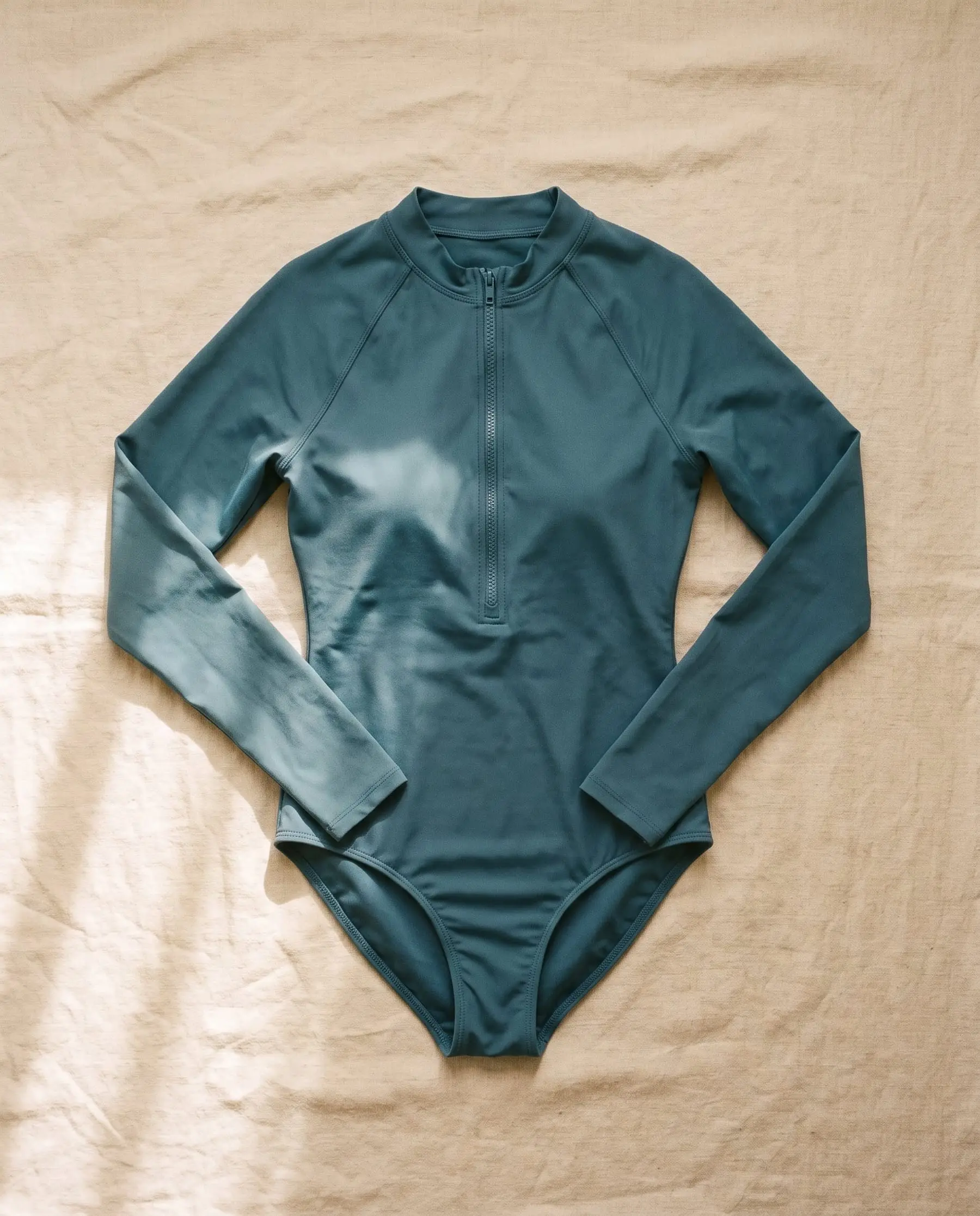 The Pass long sleeve one-piece in Tidepool, laid flat on sand-coloured linen