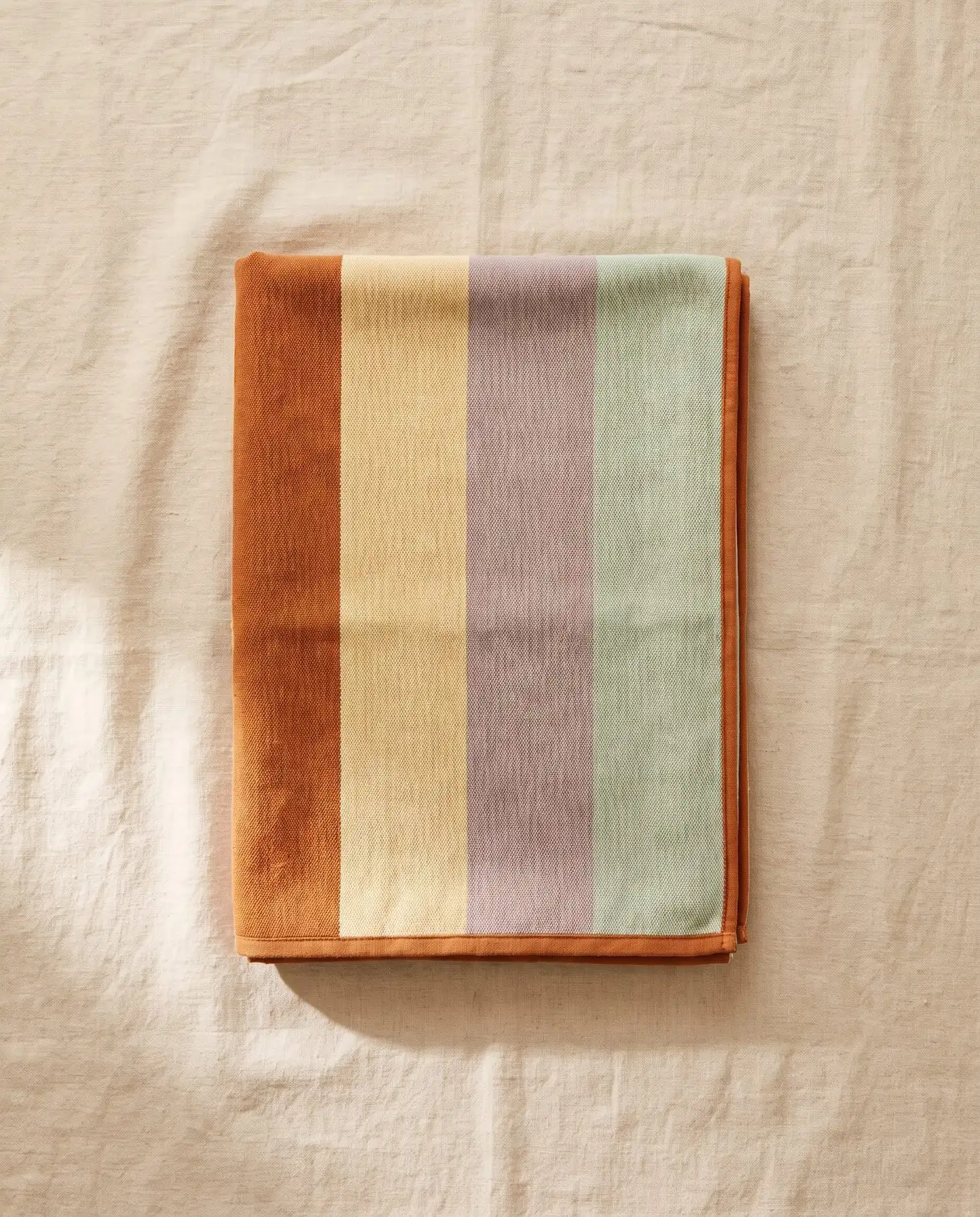 Sun-faded beach towel in wide Ochre, Driftwood and Saltbush stripes, folded on sand-coloured linen
