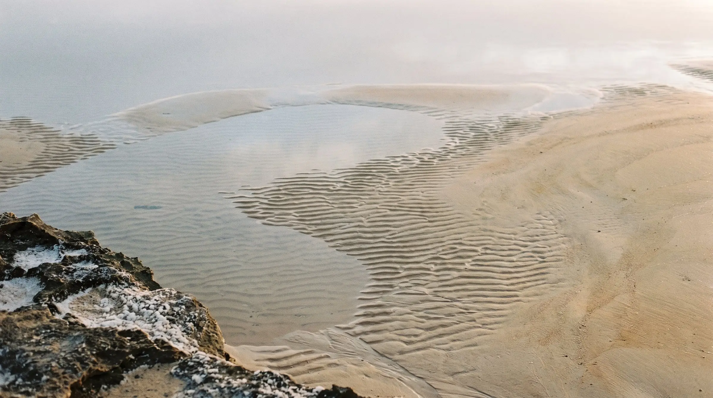 Sand ripples and a shallow tidepool at low tide beside a salt-crusted rock