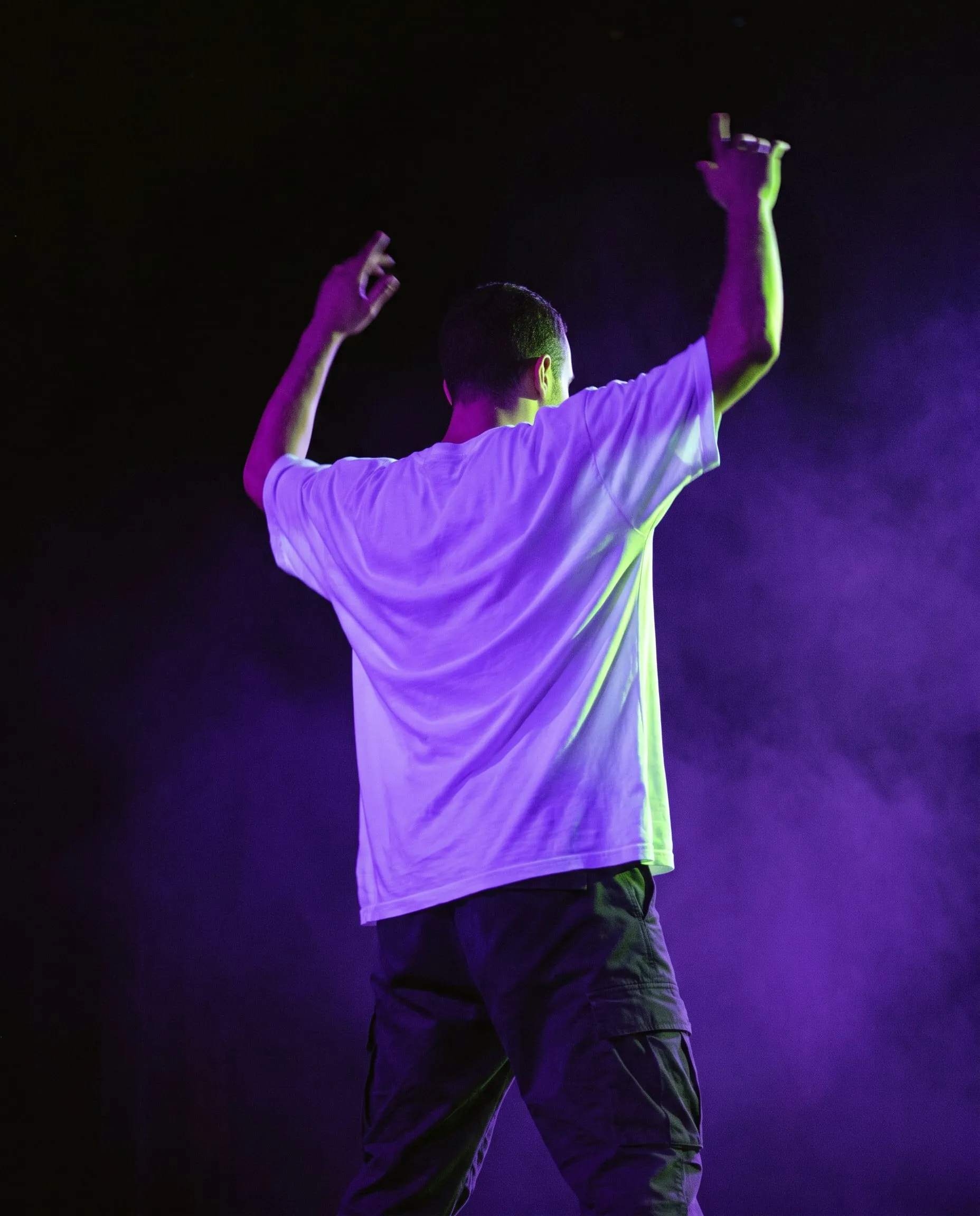 An adult dancer seen from behind with arms raised, a white t-shirt glowing in ultraviolet light