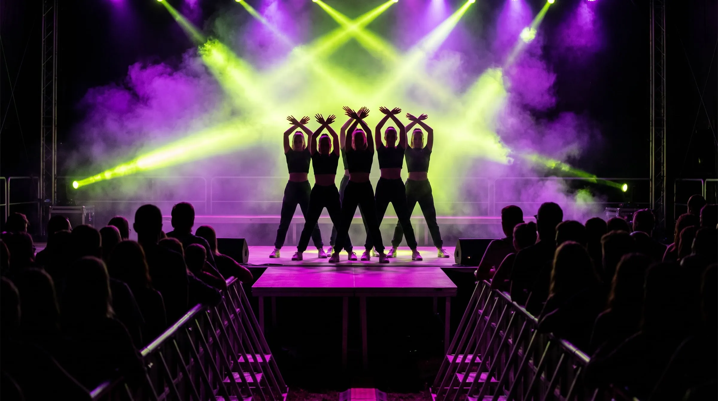 A dance crew in silhouette on an outdoor festival stage under lime and violet beams