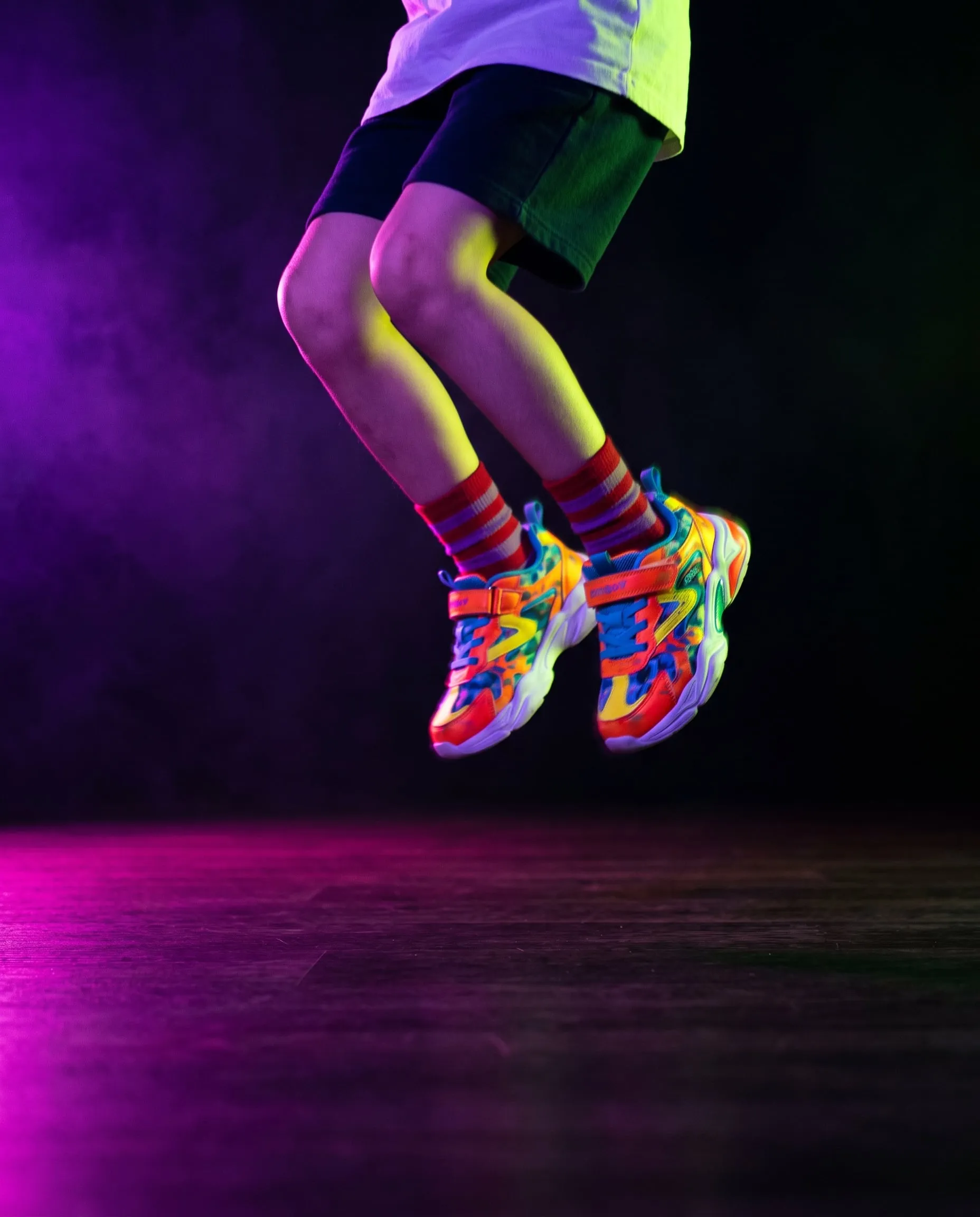 A child in bright rainbow sneakers and striped socks mid-jump under violet light