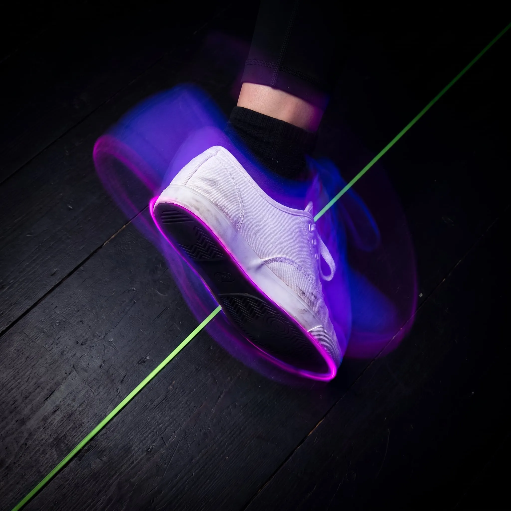 A white sneaker pivoting on a dark timber floor, blurred with speed in violet light
