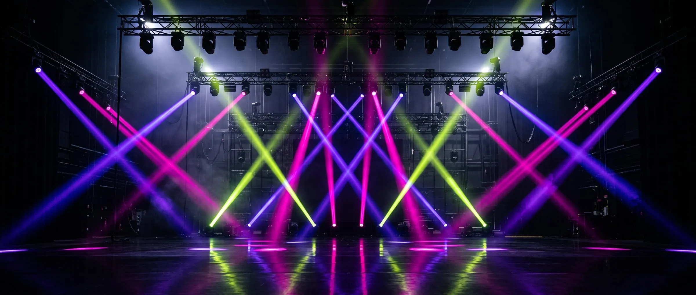 An empty stage with crossing violet, magenta and lime light beams cutting through haze