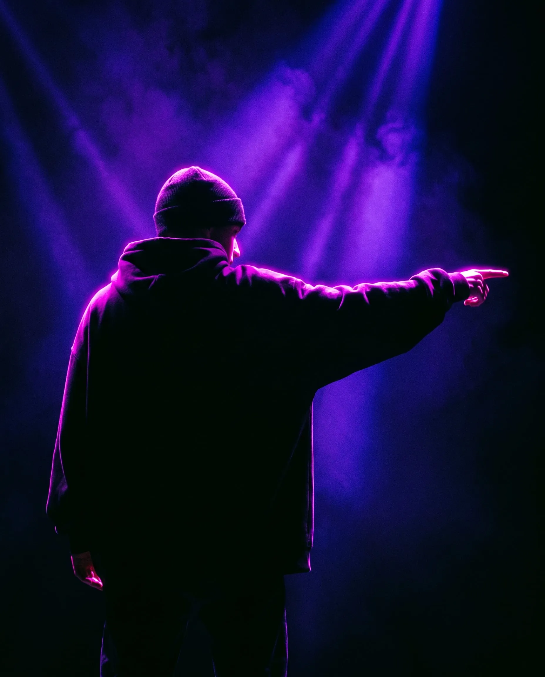 A dancer in a black hoodie seen from behind, one arm pointing, edged in magenta light