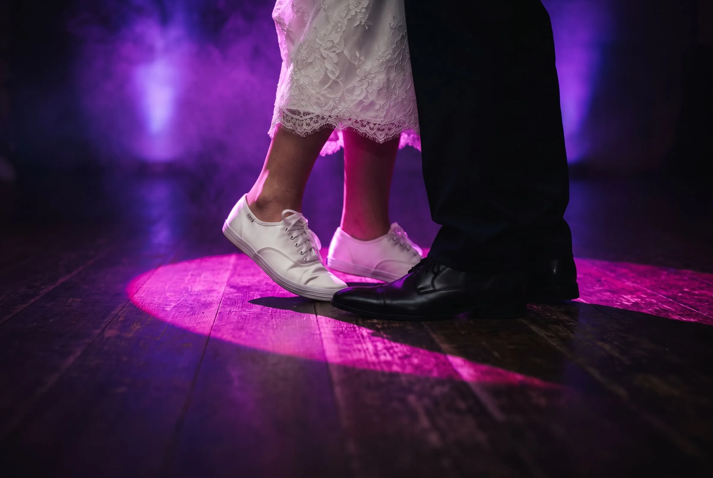 A bride in white sneakers on tiptoe beside polished black shoes in a magenta spotlight