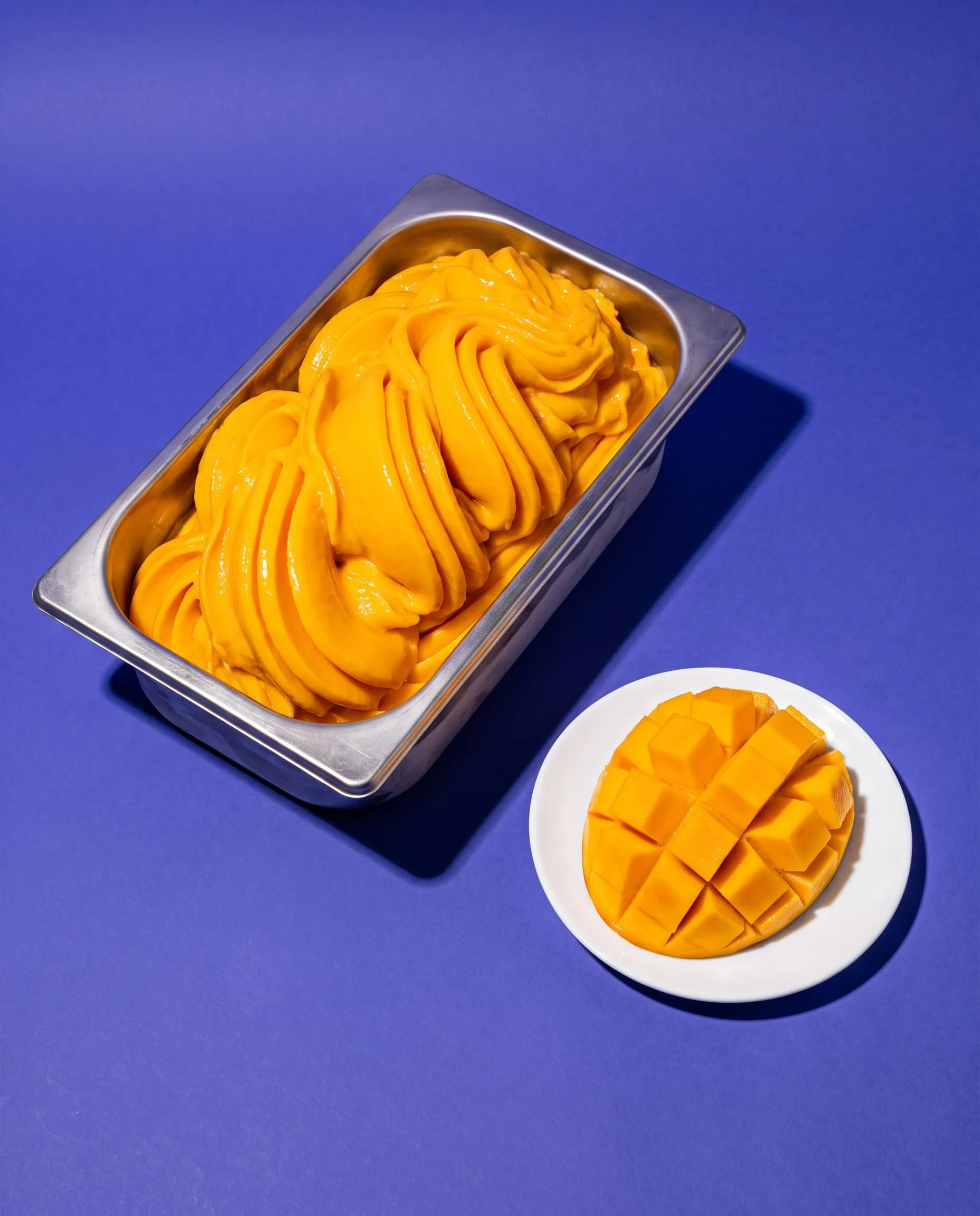 Mango sorbet swept into glossy waves in a steel tray beside a cut mango on blueberry paper