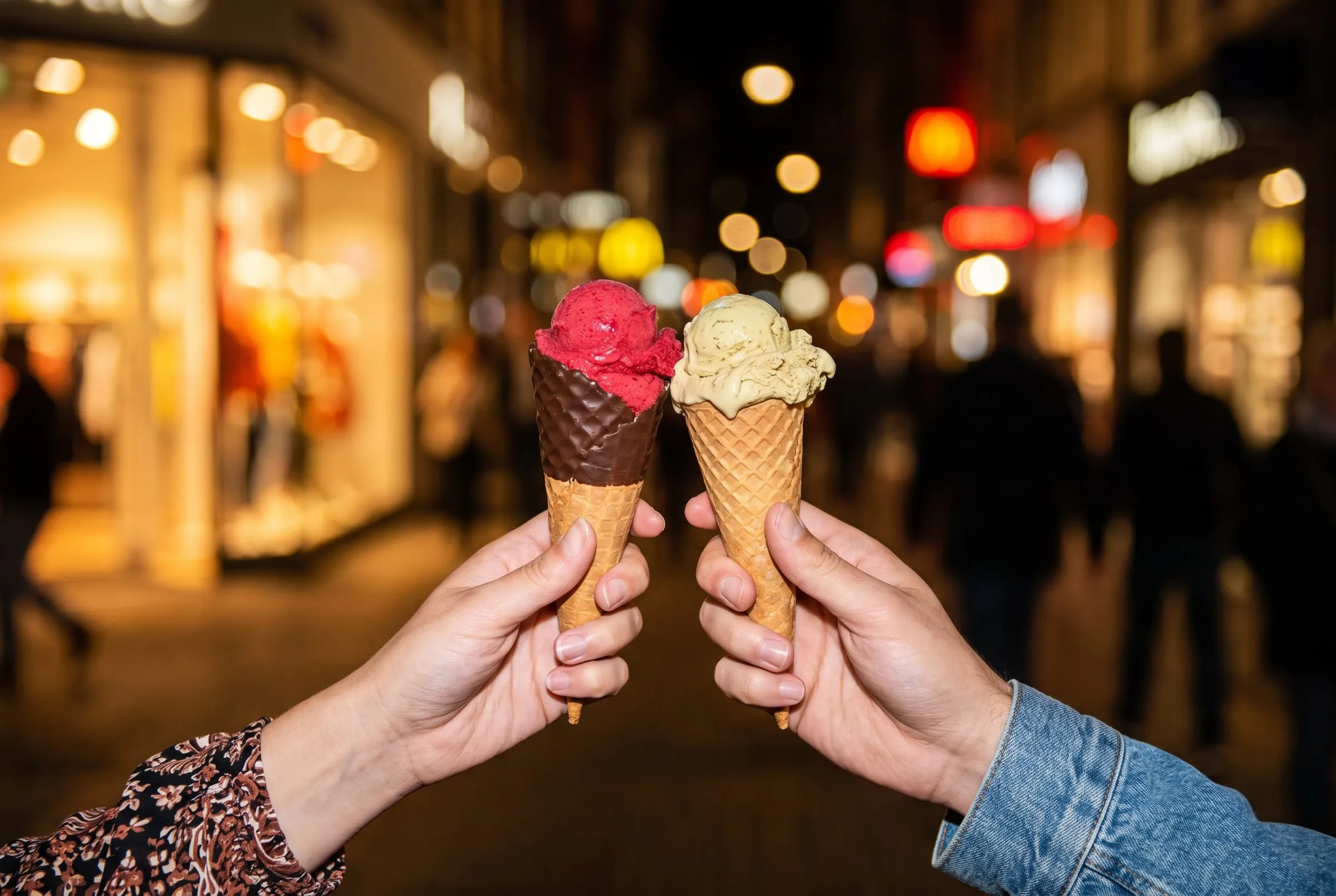 Two cones, raspberry sorbet and pistachio, clinked together on a lit-up street at night
