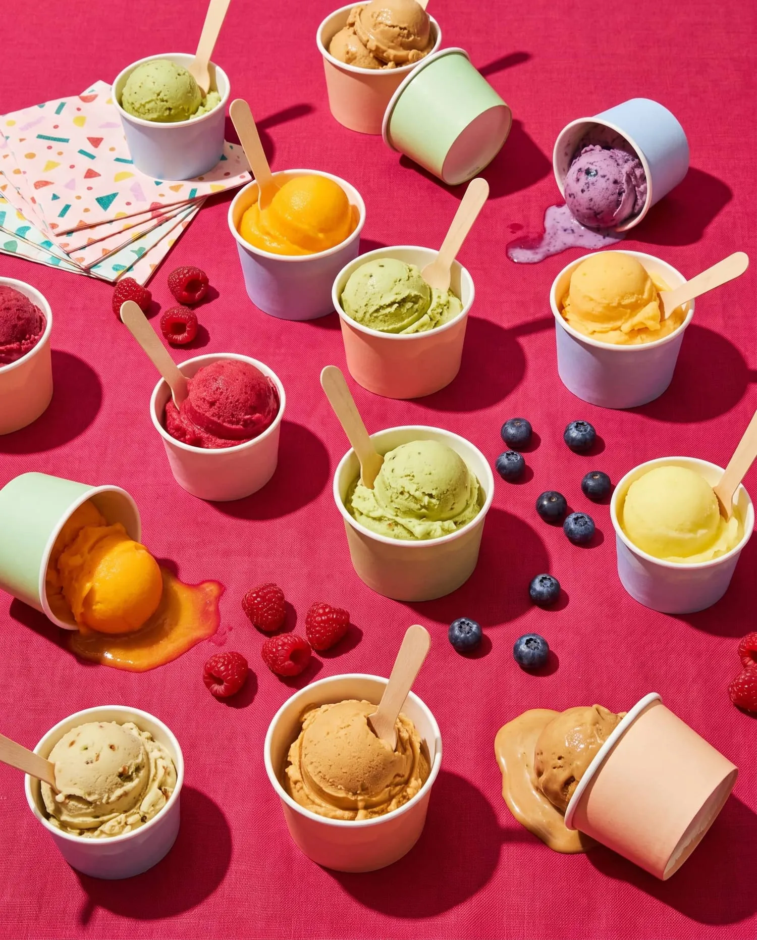 A party table on raspberry cloth crowded with cups of colourful gelato and sorbet, a few tipped over and melting