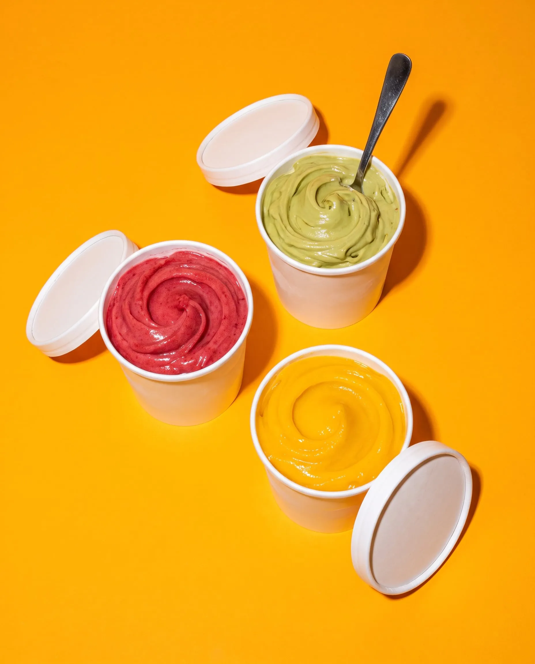Three take-home tubs of pistachio, raspberry and mango gelato with their lids off on mango orange paper