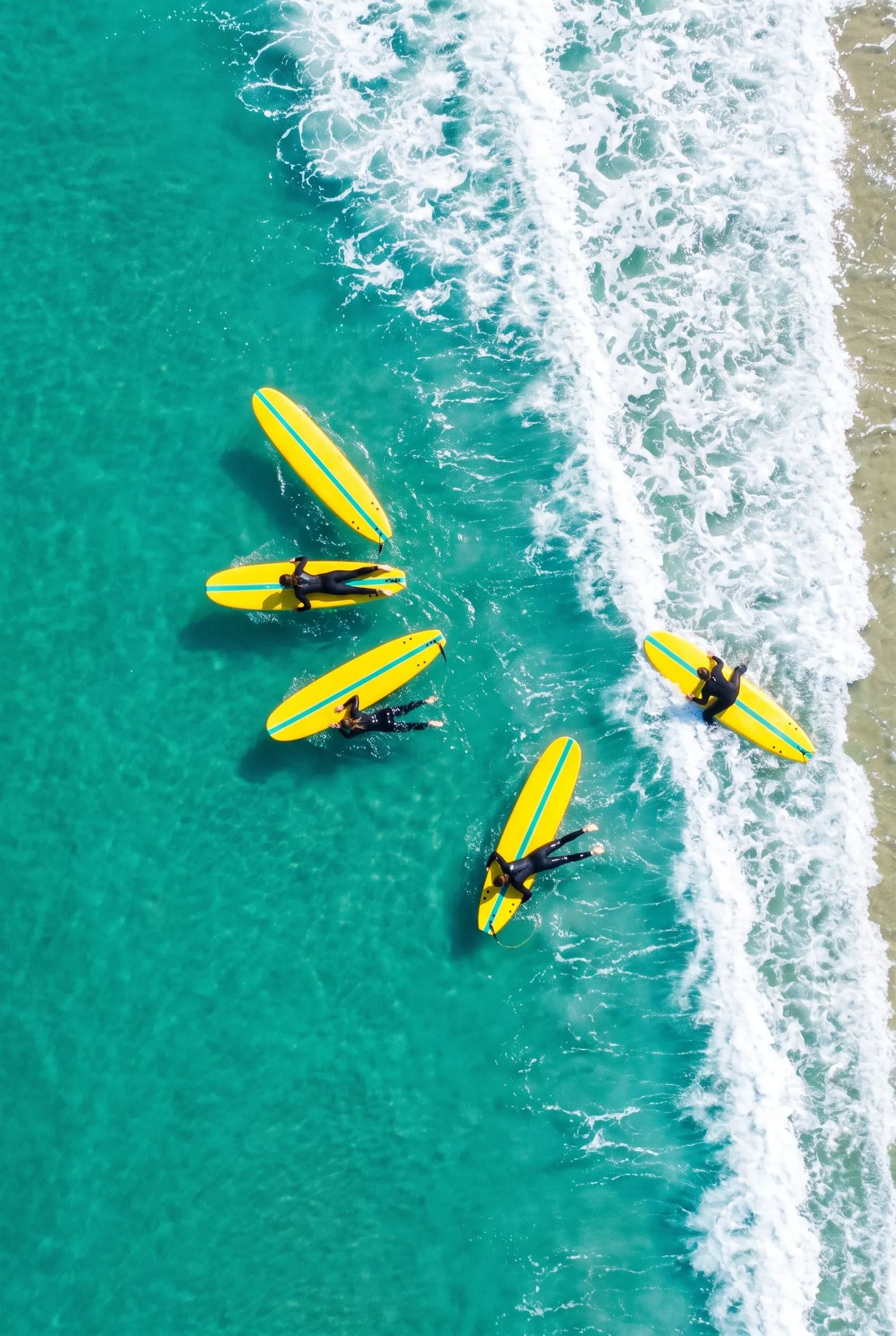 Looking straight down on turquoise water, five yellow soft-tops with learners paddling beside a line of white foam.