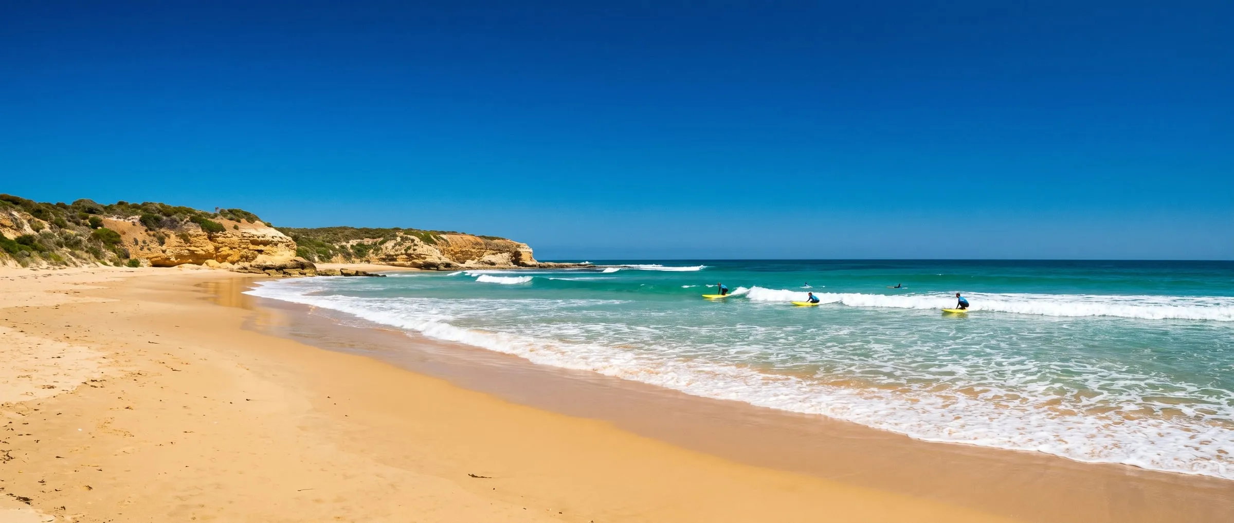 A wide Surf Coast beach with golden sand, low cliffs and small turquoise waves under a cloudless sky.