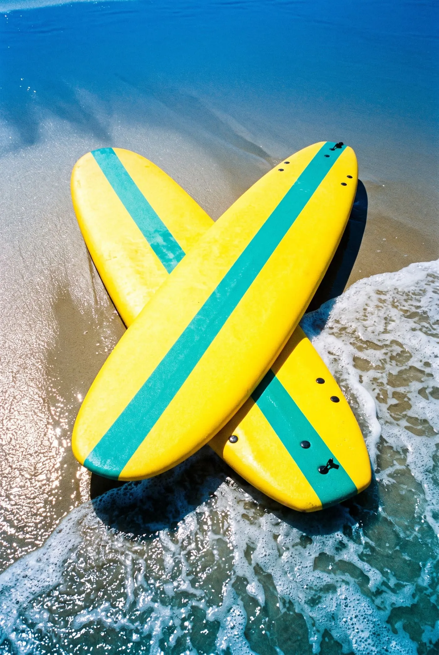 Two yellow soft-tops with teal stripes lie crossed on wet sand as a wave washes around them.