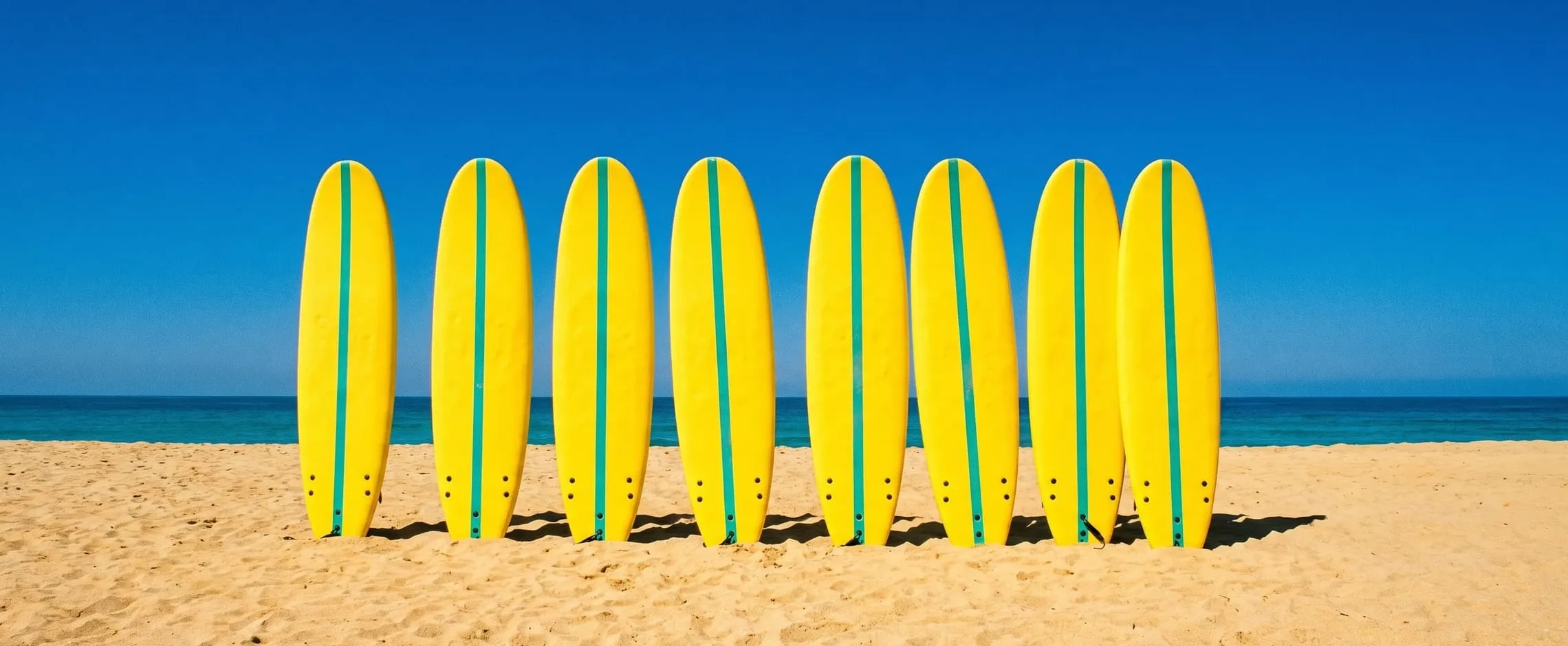 A neat row of yellow soft-tops with teal stripes standing upright in the sand under a deep blue sky.