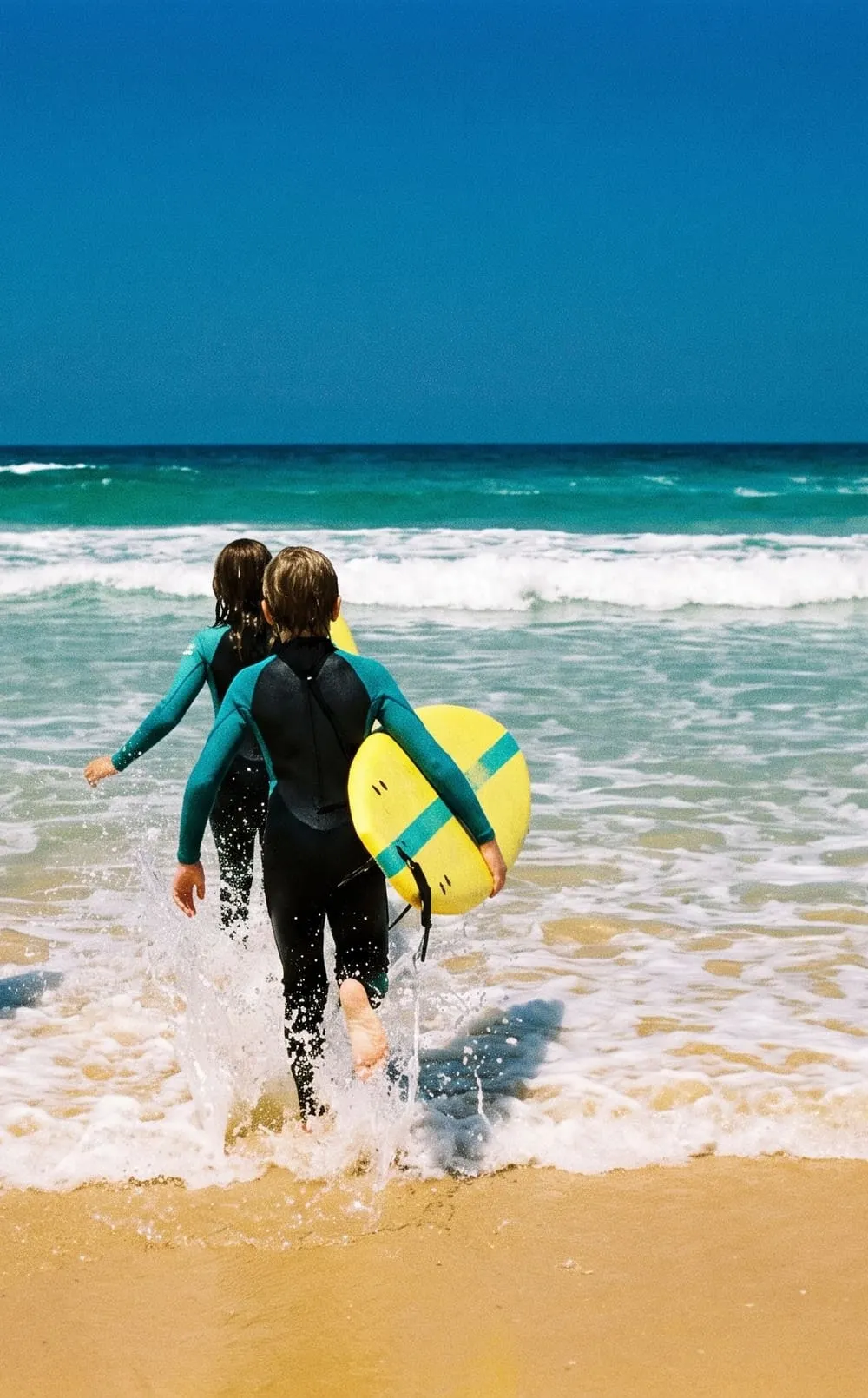 From behind, two kids in wetsuits run into the shallows carrying yellow soft-tops.