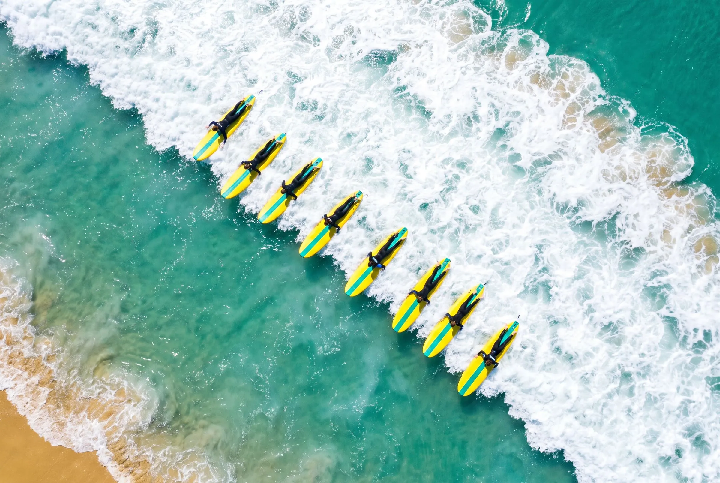 From above, a long diagonal line of learners on yellow soft-tops paddling through white foam.