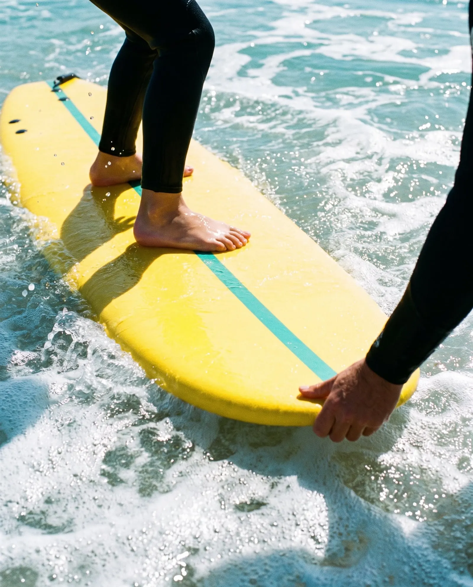 Close up of bare feet planted on a yellow soft-top in whitewater while a coach's hand steadies the tail.
