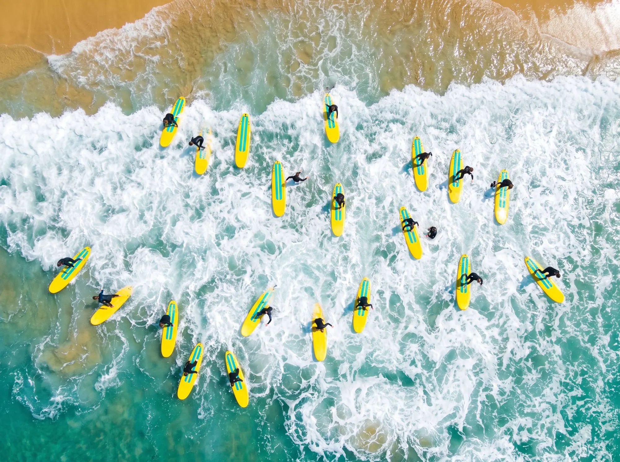 From above, a big group of learners on yellow soft-tops spread across the whitewater, some standing up, some paddling.