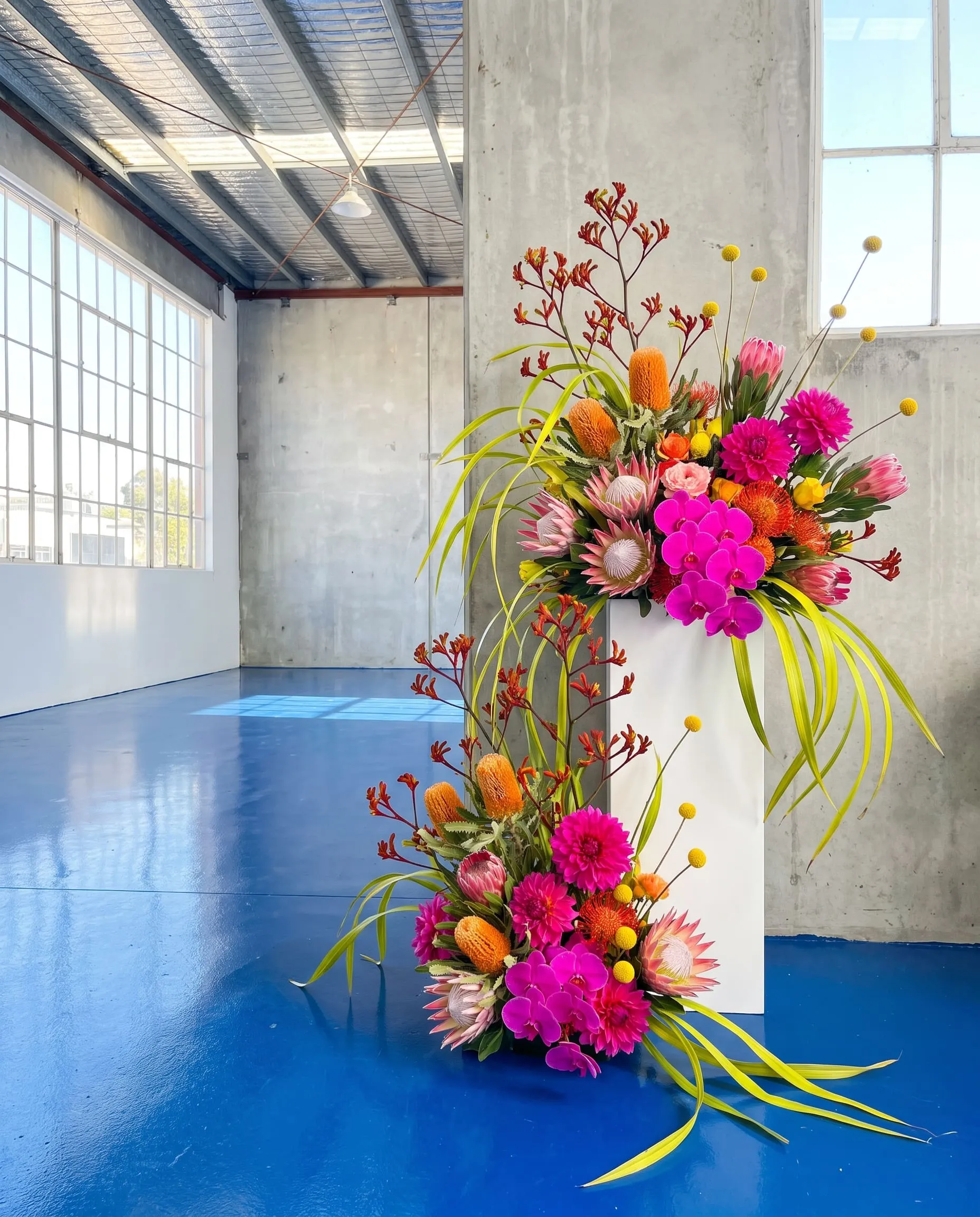 A huge, loose flower installation spilling over a white plinth in an empty, bright event space with a blue floor