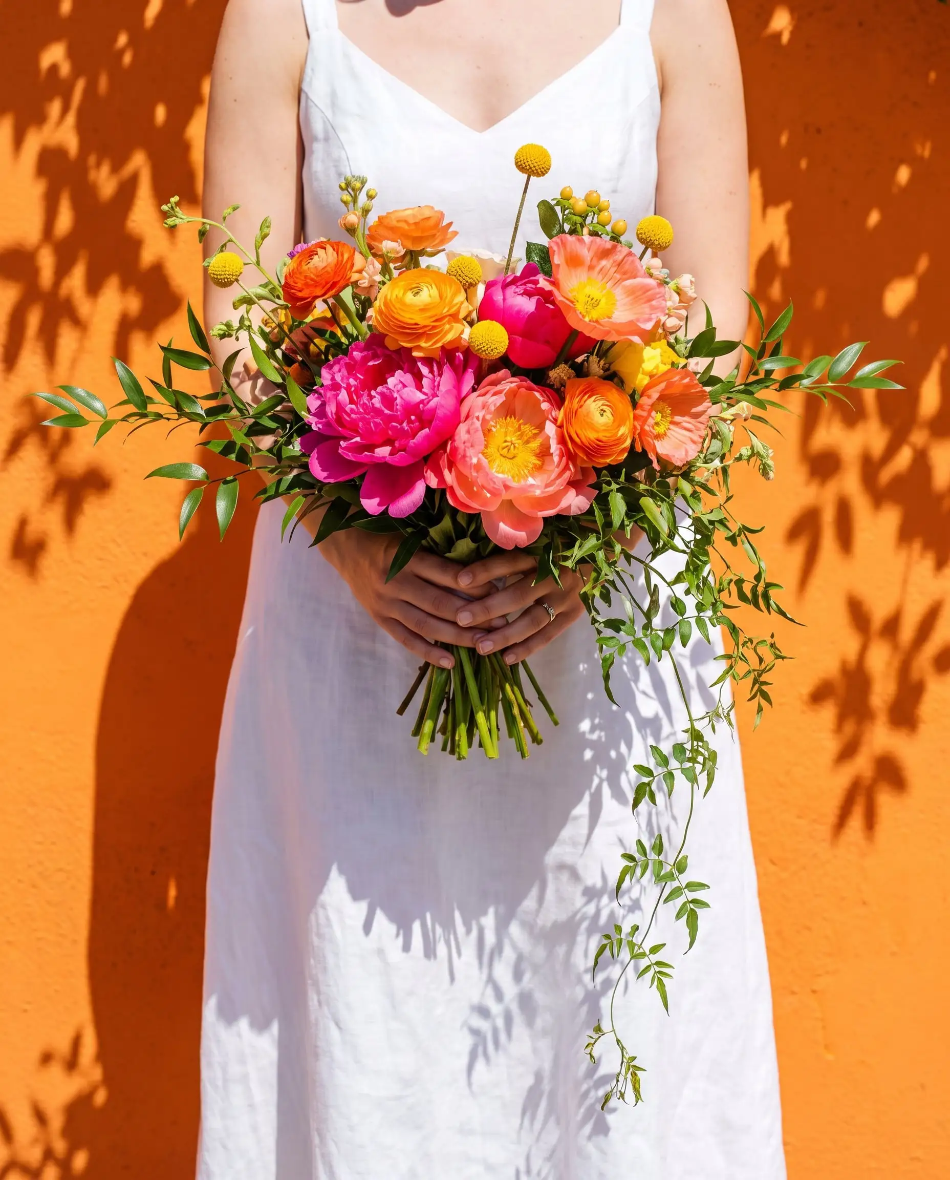Hands holding an oversized bouquet of pink peonies, orange ranunculus and coral poppies against a sunlit orange wall