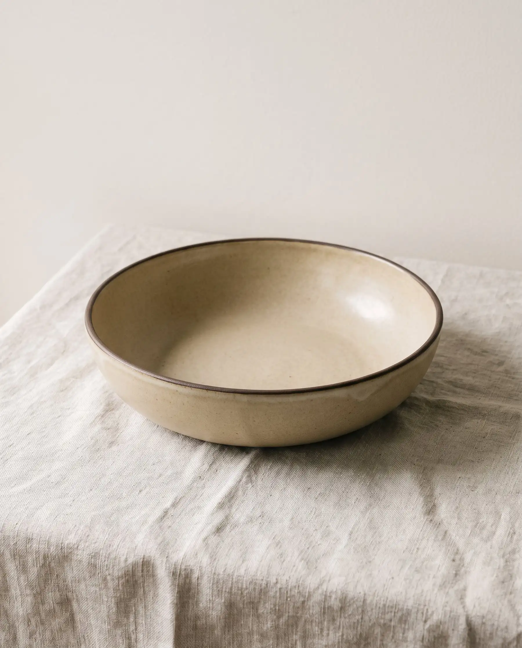 A wide shallow oatmeal glazed bowl with a dark rim on linen