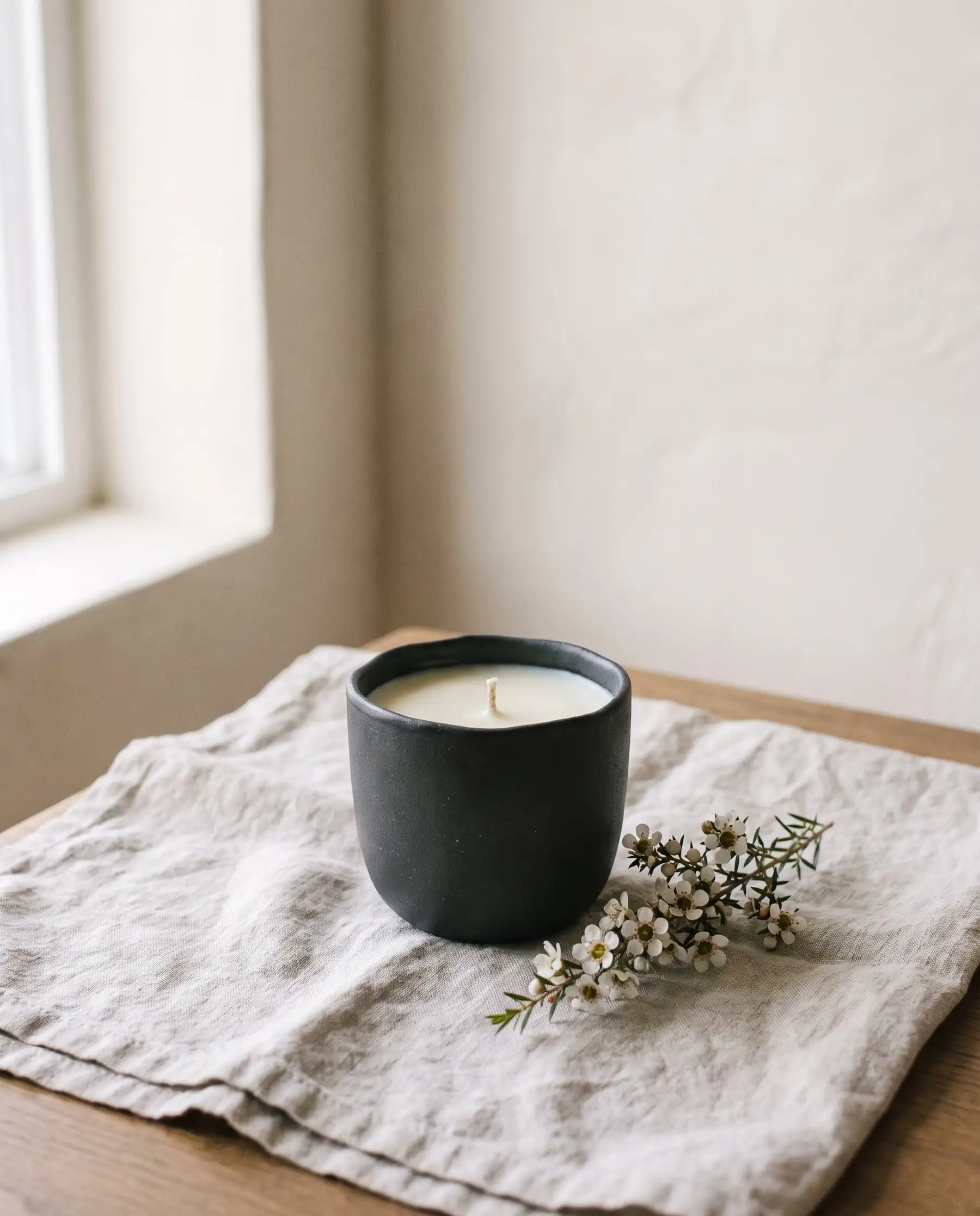 A soy candle in a matte charcoal vessel beside a sprig of tea-tree